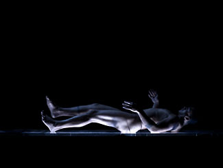 A naked body wriggles across a raised platform in spasmodic motion.